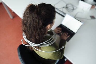 Businesswoman working on laptop tied up in cable at office - JOSEF18204