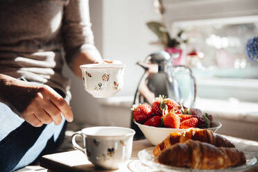 Frau hält Kaffeetasse mit Croissant und frischen Erdbeeren in der Küche - JOSEF18226