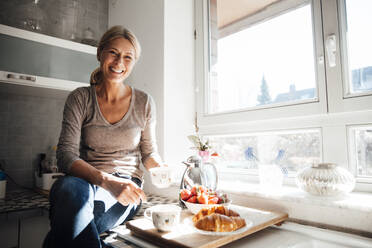 Happy woman having breakfast sitting on kitchen counter near window - JOSEF18227