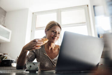 Lächelnder Freiberufler mit Croissant bei der Arbeit am Laptop im Home Office - JOSEF18228