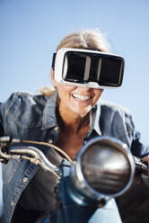 Glückliche Frau mit VR-Brille auf dem Fahrrad unter blauem Himmel - JOSEF18235