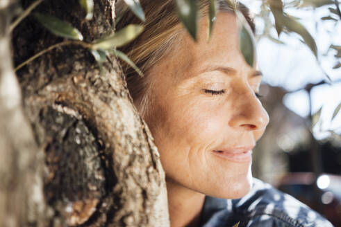 Mature woman with eyes closed embracing tree on sunny day - JOSEF18237