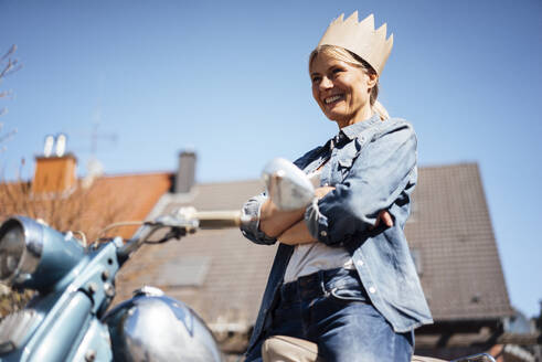 Glückliche reife Frau mit Krone steht mit verschränkten Armen auf einem Motorrad - JOSEF18240