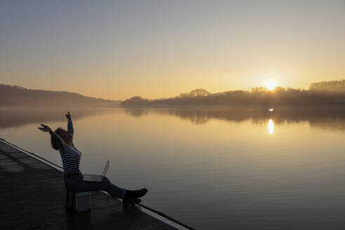 Freiberufler mit erhobenen Händen sitzt mit Laptop am See am Morgen - FLLF00870