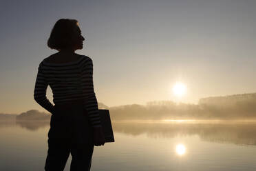 Silhouette of freelancer standing with laptop in front of lake - FLLF00872