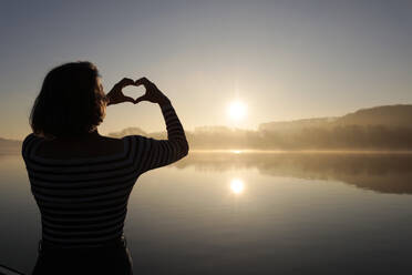 Woman making heart shape gesture in front of lake - FLLF00873