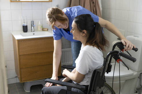 Glückliche Pflegerin im Gespräch mit einer querschnittsgelähmten Frau im häuslichen Badezimmer - MASF36119