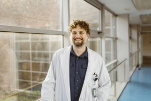 Porträt eines lächelnden reifen Arztes in einem Laborkittel, der in einem Krankenhauskorridor am Fenster steht - MASF36158