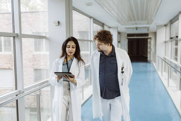 Eine Ärztin diskutiert mit einem männlichen Kollegen über ein digitales Tablet, während sie gemeinsam durch einen Krankenhausflur gehen - MASF36169