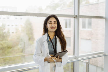Portrait of smiling female doctor holding digital tablet and pen standing against window at hospital corridor - MASF36170