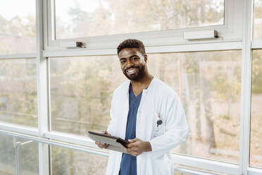 Porträt eines lächelnden jungen Arztes, der ein digitales Tablet hält und am Fenster eines Krankenhauskorridors steht - MASF36171