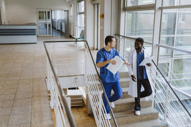Männlicher Arzt und Krankenschwester diskutieren in voller Länge über Dokumente, während sie auf einer Treppe im Krankenhaus nach unten gehen - MASF36183