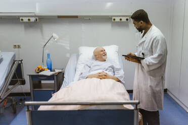 Junger männlicher Arzt mit digitalem Tablet im Gespräch mit einem älteren Patienten, der auf einem Bett im Krankenhaus liegt - MASF36200