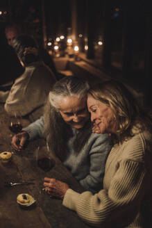 Lächelnde ältere weibliche Freunde mit Weingläsern, die während einer Party am Esstisch sitzen - MASF36231