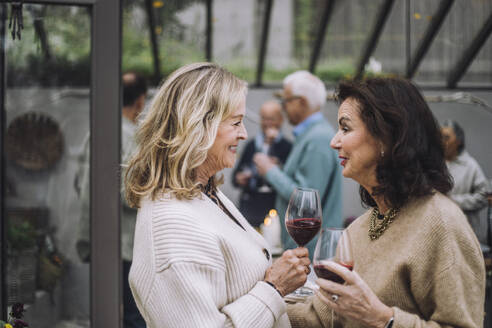 Lächelnde ältere Frau mit Weingläsern im Gespräch mit Freundinnen bei einer Dinnerparty - MASF36243