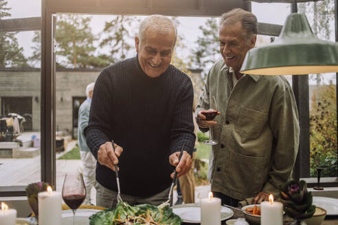 Glücklicher reifer Mann, der mit einem männlichen älteren Freund auf einer Dinnerparty steht und Salat zubereitet - MASF36266