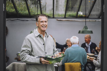 Porträt eines lächelnden älteren Mannes, der eine Salatschüssel bei einer Dinnerparty hält - MASF36279