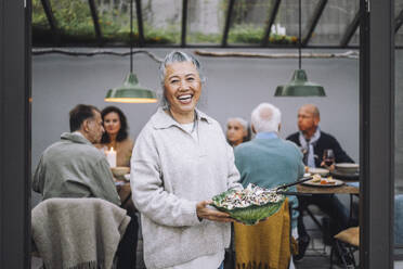 Porträt einer fröhlichen älteren Frau mit einer Salatschüssel in der Hand bei einer Dinnerparty - MASF36280