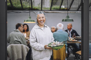 Porträt einer fröhlichen älteren Frau mit einer Salatschüssel in der Hand bei einer Dinnerparty - MASF36280