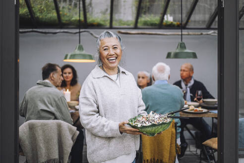 Portrait of cheerful senior woman holding salad bowl at dinner party - MASF36280