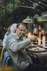 Smiling senior man holding wineglass while sitting at dining table during party - MASF36287