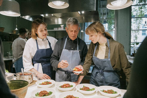 Chefkoch instruiert männliche und weibliche Schüler beim Anrichten während eines Kochkurses in der Küche - MASF36325