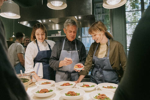 Lächelnde Köchin mit Schülern, die während eines Kochkurses in der Küche Teller anrichten - MASF36326