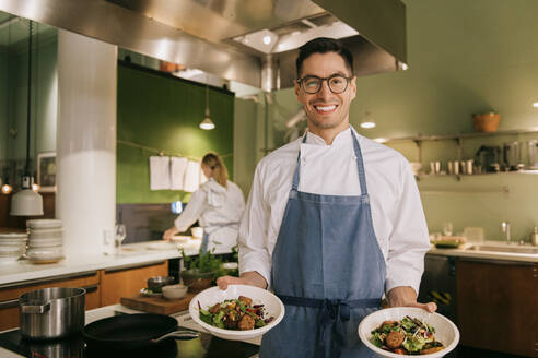 Portrait of confident male chef showing food while standing in commercial kitchen - MASF36351