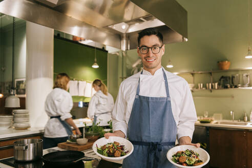 Lächelnder männlicher Koch, der Schüsseln mit Lebensmitteln hält, während er in einer Restaurantküche steht - MASF36352