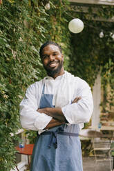 Porträt eines selbstbewussten männlichen Kochs, der mit verschränkten Armen vor einem Restaurant steht - MASF36355