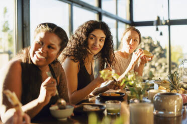 Lächelnde Freundinnen schauen weg, während sie beim Frühstück am Esstisch sitzen - MASF36466