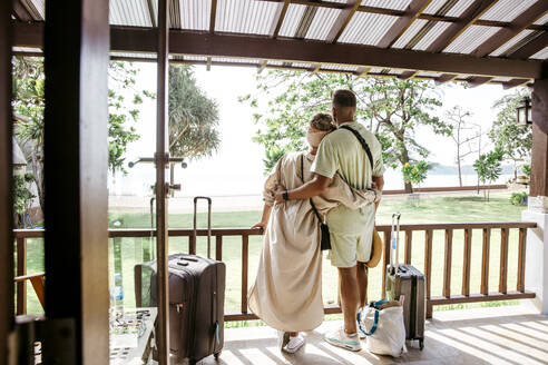 Rear view of male and female tourists with arms around standing on porch of villa - MASF36532