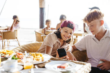 Glückliches Mädchen, das sein Smartphone mit seinem Bruder in einem Ferienort teilt - MASF36560