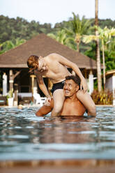 Cheerful father carrying son on shoulders in swimming pool at resort during vacation - MASF36566