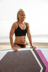 Smiling woman with paddleboard standing against clear sky at beach - MASF36589