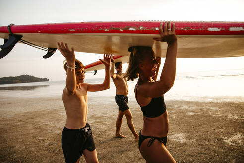 Happy family carrying paddleboards on heads while walking at beach during sunset - MASF36593