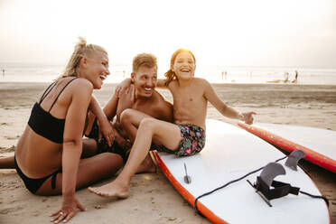 Fröhliche Eltern genießen mit Tochter sitzen auf Paddleboard am Strand bei Sonnenuntergang - MASF36599