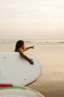 Mädchen mit Paddleboard zeigt auf das Meer am Strand bei Sonnenuntergang - MASF36602