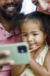 Nettes Mädchen, das mit seinen Eltern zu Hause auf das Smartphone schaut - IKF00037