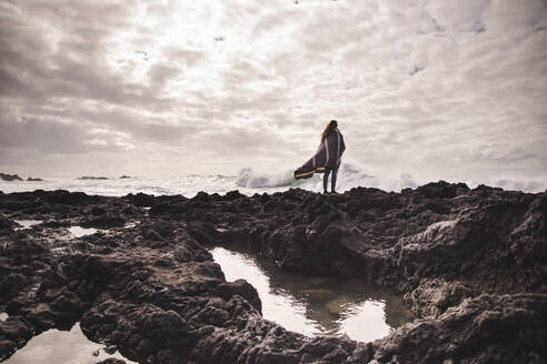 Junge Frau im Poncho steht auf einem Felsen vor dem Meer - PCLF00346