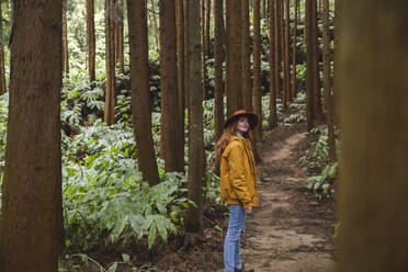 Rothaarige Frau mit Hut steht inmitten hoher Bäume im Wald - PCLF00357