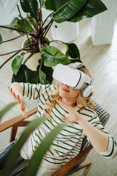 Smiling woman playing games wearing VR glasses sitting in chair at home - MDOF00981