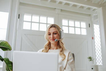 Lächelnder Freiberufler mit Headset bei der Arbeit am Laptop zu Hause - MDOF00994