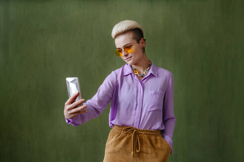Eine stilvolle junge Frau macht ein Selfie vor einer üppigen grünen Wand und trägt dabei eine trendige Sonnenbrille - YTF00708