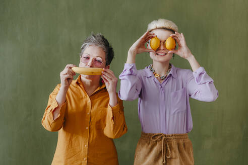 Reife Frau hält Banane mit Tochter bedeckt Augen mit Zitronen vor der Wand - YTF00713