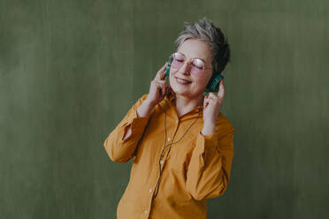 Lächelnde Frau mit geschlossenen Augen, die über Kopfhörer vor einer grünen Wand Musik hört - YTF00727