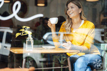 Happy woman holding mobile phone with coffee cup sitting at table in cafe - JOSEF18253