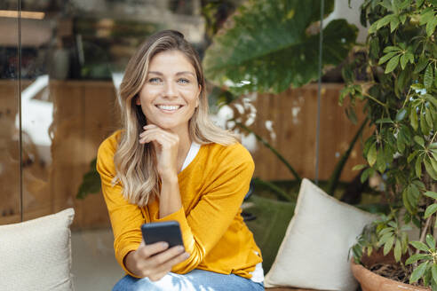 Glückliche Frau, die ein Smartphone hält und mit der Hand am Kinn neben einer Pflanze im Café sitzt - JOSEF18269