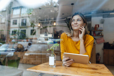 Lächelnde Frau mit Tablet-PC sitzt am Tisch in einem Café durch Glas gesehen - JOSEF18283