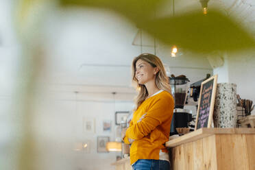 Thoughtful smiling woman standing with arms crossed in cafe - JOSEF18303